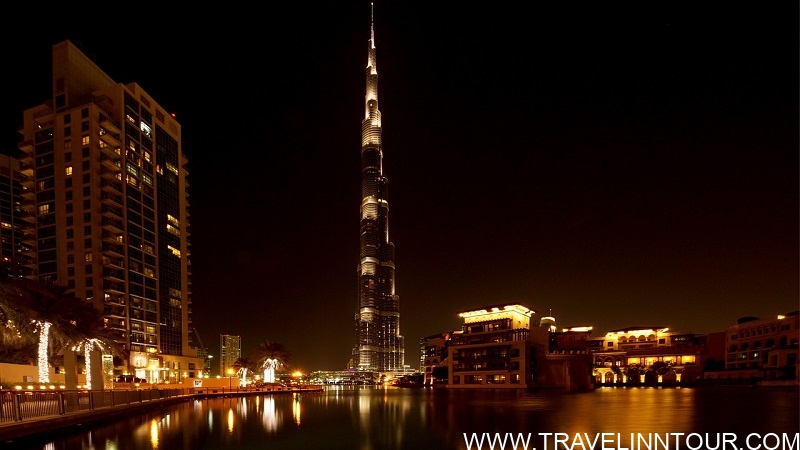 Night view of the illuminated Burj Khalifa towering over Dubai's cityscape, surrounded by other buildings and a calm waterway reflecting the lights, showcasing Dubai's vibrant nightlife and iconic architecture.