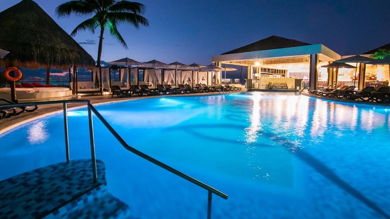 Luxurious resort pool area at night with illuminated swimming pool, palm trees, and cabanas, suitable for travelers seeking clothing optional vacations and nudist hotel experience