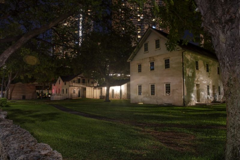 Night view of historic missionary buildings in Hawaii, surrounded by lush greenery and illuminated by soft lighting. The city skyline is visible in the background, creating a blend of traditional and modern elements, perfect for exploring Hawaii's cultural heritage.