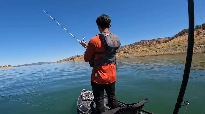 Angler wearing an orange shirt and life jacket fishing from a kayak on a clear, calm lake in California, with distant hills and clear blue sky, ideal for bass fishing.