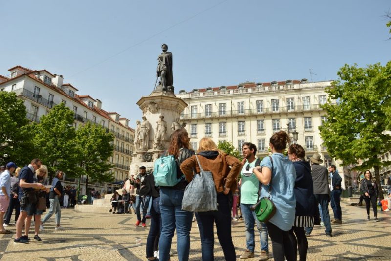 Tourists gather around the Pedro IV Square statue in Lisbon, enjoying a guided walking tour under a sunny sky, exploring the city's historical and architectural highlights.