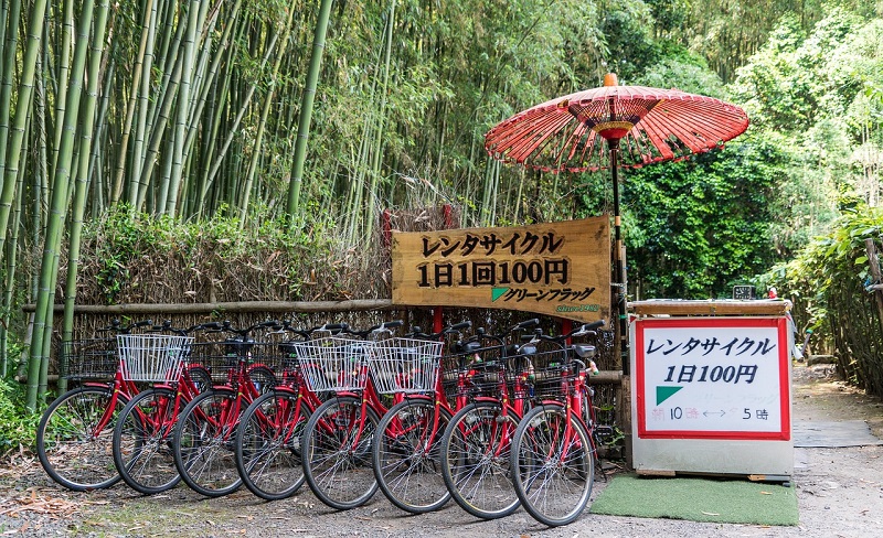 Red rental bicycles lined up in front of a bamboo grove with a traditional red umbrella and wooden sign offering rentals for 100 yen per day.