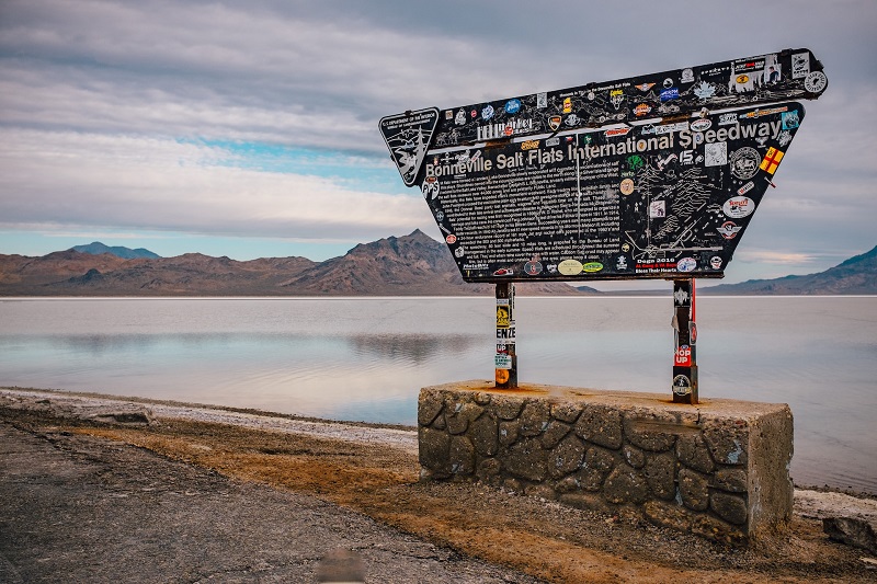 Signage at Bonneville Salt Flats International Speedway with a scenic view of vast salt flats and mountains in the background, under a partly cloudy sky in Utah.