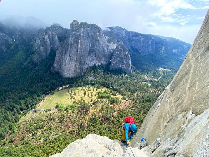 Climber ascending a steep rock face in Yosemite National Park, with a panoramic view of lush green forests and towering granite cliffs under a cloudy sky.