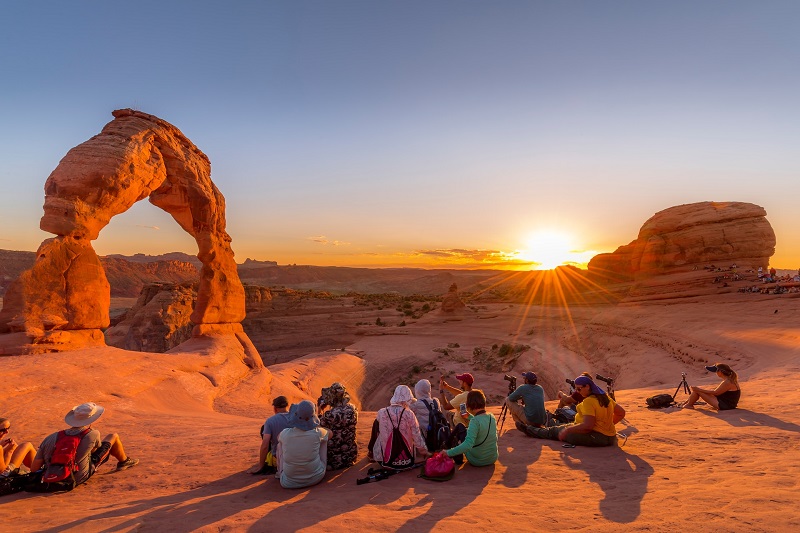 A group of people enjoying the sunset at Delicate Arch in Arches National Park, Utah. The iconic arch is bathed in golden light, highlighting the stunning red rock formations and natural beauty of the landscape, perfect for nature lovers and outdoor adventurers.