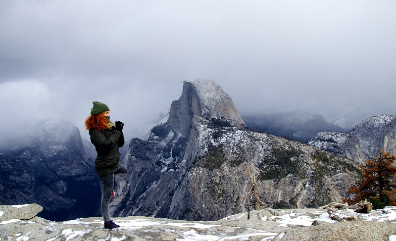 Person practicing yoga pose on a snowy cliff with a view of Half Dome in Yosemite National Park.