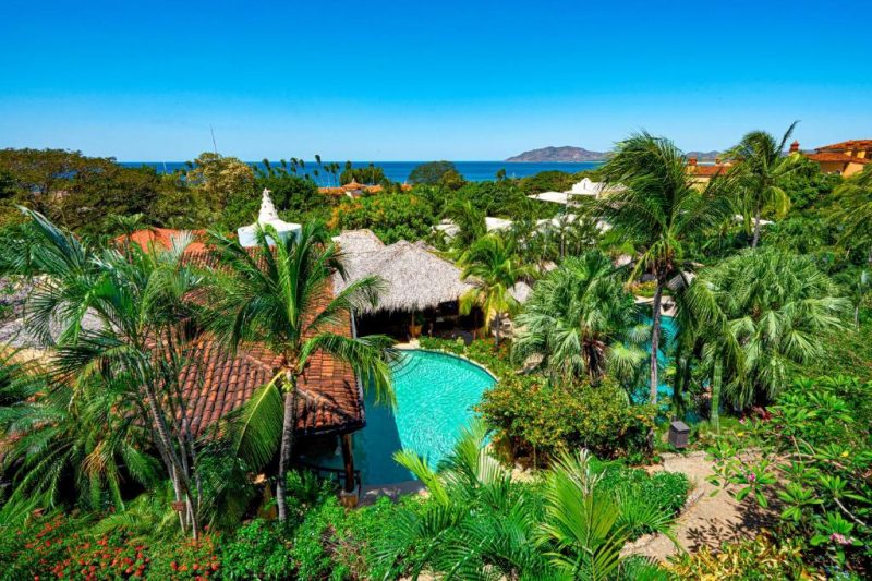 Panoramic view of a tropical resort in Costa Rica featuring lush greenery, a pool, thatched-roof structures, and ocean in the background, ideal for adults and naturists.