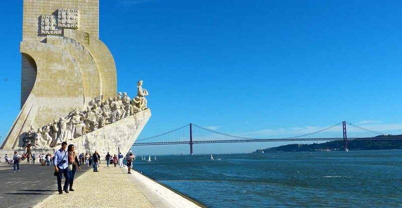 Monument of Discoveries in Lisbon with people walking along the riverside path, showcasing the 25th of April Bridge in the background on a clear, sunny day.