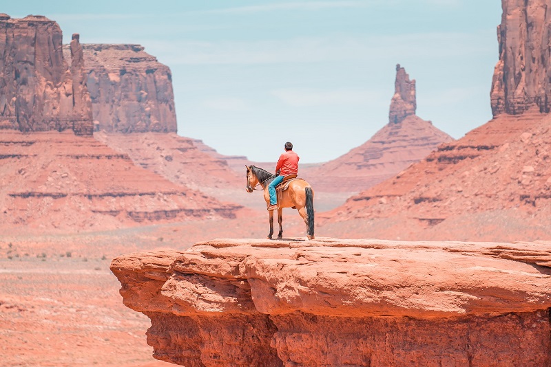 Person on horseback on a red rock cliff in Utah's Monument Valley, surrounded by dramatic sandstone formations under a clear blue sky, perfect for adventurous nature lovers.