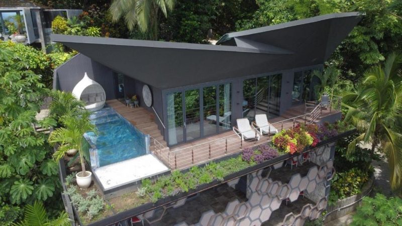 Aerial view of a modern, luxury villa in Costa Rica surrounded by lush greenery. The villa features a sleek angular design, a private pool, and a spacious wooden deck with lounge chairs, ideal for a relaxing and private getaway at a clothing optional resort for adults and naturists.