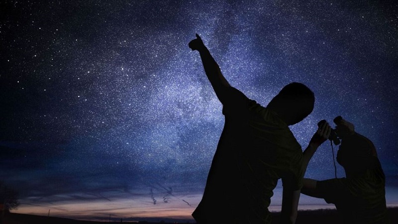 Silhouette of two people stargazing, one pointing at the Milky Way in a clear night sky, symbolizing outdoor adventures in Utah.