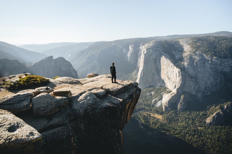 Person standing on a rocky cliff in Yosemite National Park, overlooking vast mountain landscape and valley below