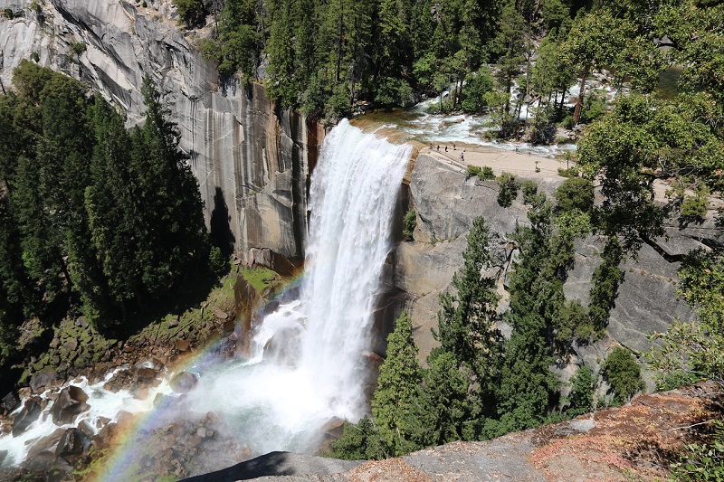 A breathtaking view of Vernal Fall in Yosemite National Park, surrounded by lush green trees and granite cliffs, with a vivid rainbow at the base of the waterfall.