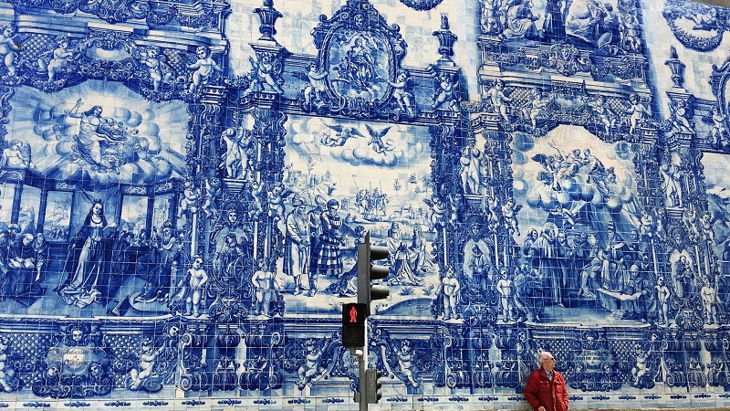 Azulejos tiles mural in Lisbon depicting historical scenes in blue and white, with a pedestrian and traffic light in the foreground.