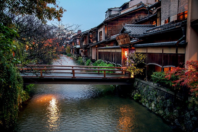 Traditional wooden houses line a serene canal with a charming wooden bridge in Kyoto, Japan, during twilight, highlighting the city's historic architecture and tranquil atmosphere.