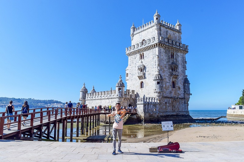 Violinist performing in front of the iconic Belem Tower in Lisbon, Portugal, with clear blue skies and a scenic view, highlighting a popular stop on walking tours.