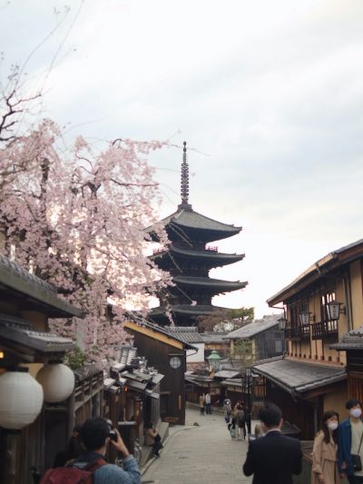 Traditional Kyoto street with people walking, featuring a pagoda and cherry blossom trees, representing Japanese culture and architecture.
