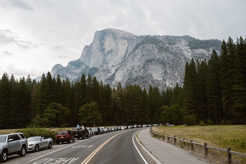 Scenic view of Half Dome in Yosemite National Park with a line of cars on the road, surrounded by tall pine trees and cloudy sky.