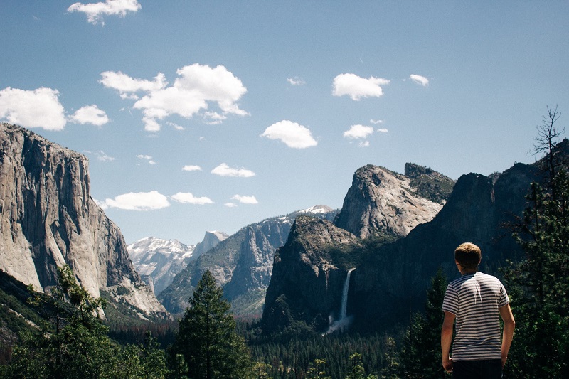 Scenic view of Yosemite National Park with person admiring the landscape, featuring majestic mountains, lush greenery, and a waterfall under a clear blue sky.