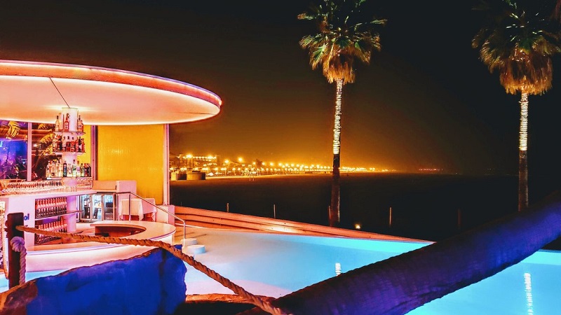 Valencia beachside bar at night with illuminated pool, palm trees, and city lights in the background, highlighting nightlife and travel attractions.