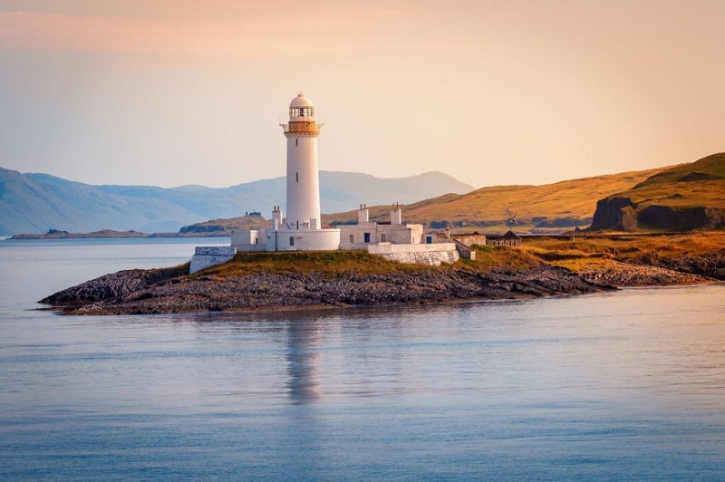 Scenic view of a lighthouse on a small island off the coast of Scotland at sunset, surrounded by calm waters and distant hills, showcasing the natural beauty ideal for a perfect vacation.