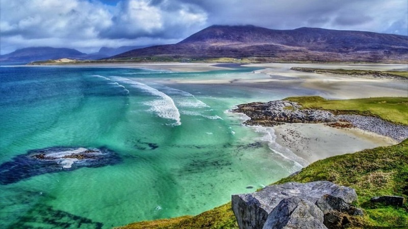 Scenic view of a pristine beach with turquoise waters and green cliffs on a Scottish island under a cloudy sky, ideal for a perfect vacation getaway.