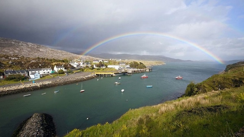 Scenic view of a coastal village in Scotland under a vibrant rainbow, with boats in a calm harbor and green hills in the background.