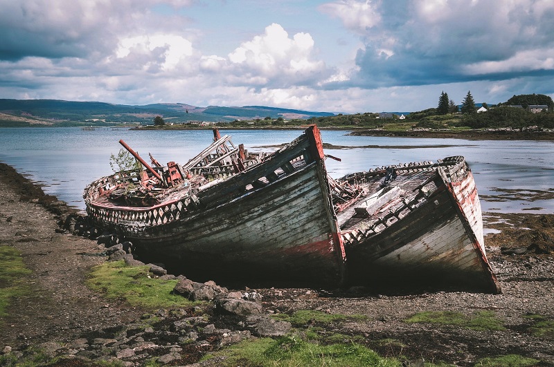 Rustic abandoned boats lying on the shore of a scenic Scottish island, surrounded by calm waters and lush greenery under a partly cloudy sky, capturing the tranquil and historic essence of Scottish island landscapes.
