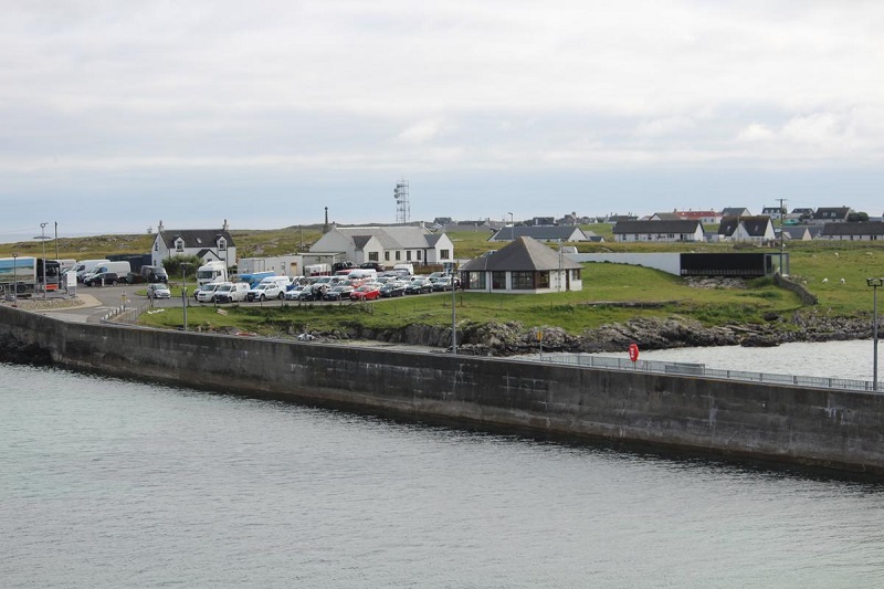 Scottish island harbor view with cars parked, buildings, and a grassy area, under a cloudy sky, ideal for exploring top vacation destinations.