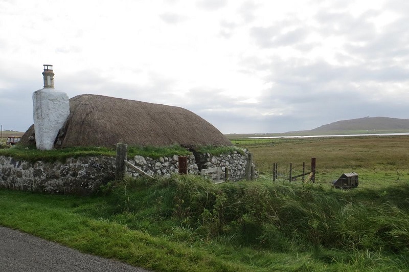 A traditional Scottish blackhouse with a thatched roof and stone walls, set in a grassy and rural landscape on a cloudy day.
