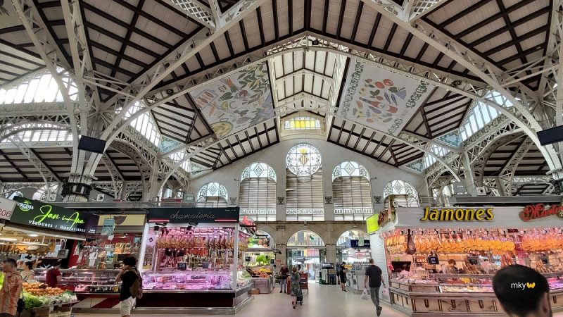 Interior view of Valencia's central market showcasing bustling food stalls with cured meats and produce, under a beautifully designed ceiling with stained glass windows.