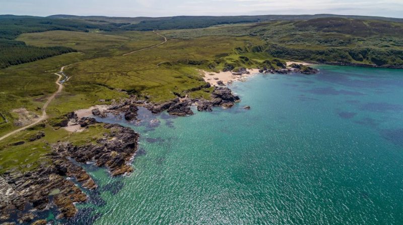 Aerial view of a picturesque Scottish island coastline featuring rugged cliffs, lush green landscape, and turquoise waters under a clear sky, perfect for a vacation exploring top islands in Scotland.