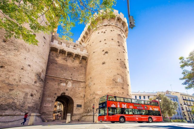 A red double-decker sightseeing bus in front of the historic Torres de Serranos gate in Valencia under a clear blue sky, showcasing a popular city attraction.