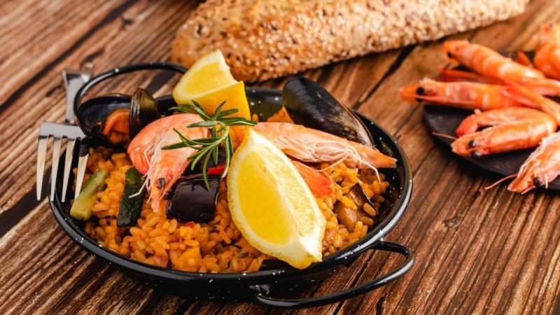 Plato de paella valenciana con mariscos, rodajas de lim&oacute;n y pan de semillas sobre una mesa de madera, representando la cocina tradicional de Valencia.