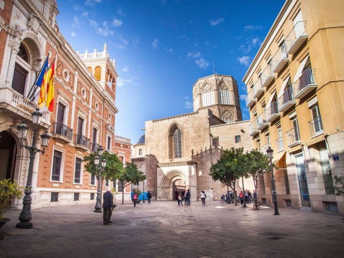 Plaza in Valencia with historic architecture, featuring the Valencia Cathedral and government buildings decorated with flags, under a clear blue sky.