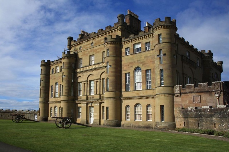 Culzean Castle, a large sandstone structure with turrets and battlements, stands majestically against a blue sky on a lush green lawn, symbolizing Scotland's rich heritage and history.