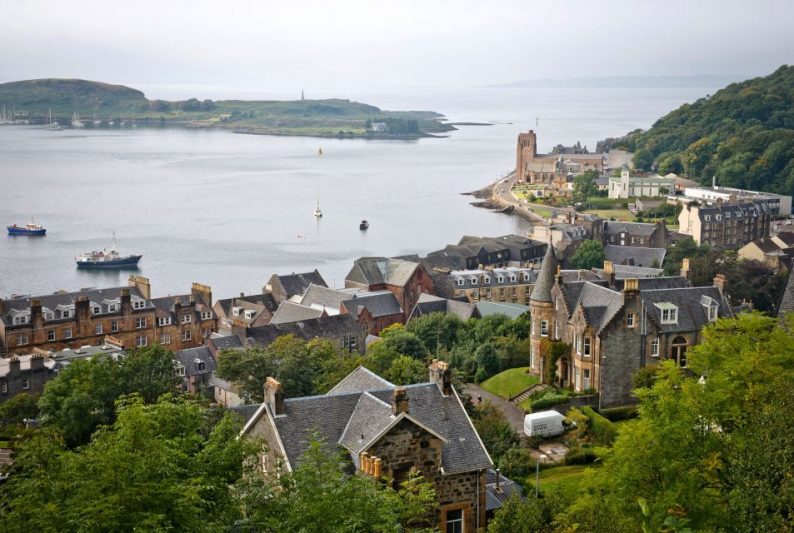 View of a picturesque coastal town in Scotland with traditional stone buildings overlooking a serene bay. Boats are anchored in the water, and lush greenery surrounds the area, highlighting the scenic beauty ideal for a perfect island vacation.