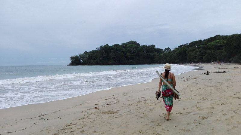 Person walking on a secluded beach in Costa Rica, carrying a surfboard and wearing tropical attire on a serene sandy shore with lush green trees in the background.