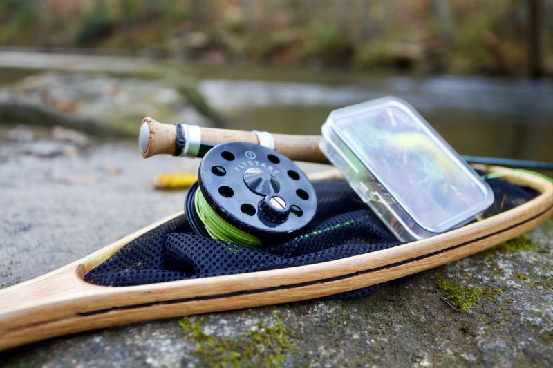 Fly fishing gear including a fly reel, wooden net, and a box of flies placed on a rock by a Canadian lake, highlighting fishing equipment for top fishing spots in Canada.