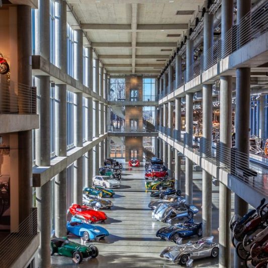 Interior view of a spacious car museum in Texas, showcasing an extensive collection of classic and vintage cars arranged in rows. The high-ceilinged building features large windows and structural beams, enhancing the display of automotive history.