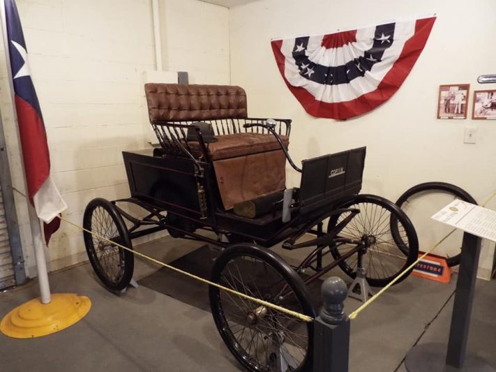 Vintage car exhibit in a Texas museum showcasing early automotive history with a classic open-top carriage car, surrounded by a Texas flag and patriotic decor.