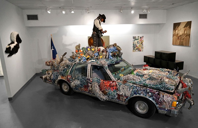 An intricately decorated car with colorful mosaic and floral designs on display in a museum room, featuring a person dressed in cowboy attire standing on the roof, embracing Texan art culture.