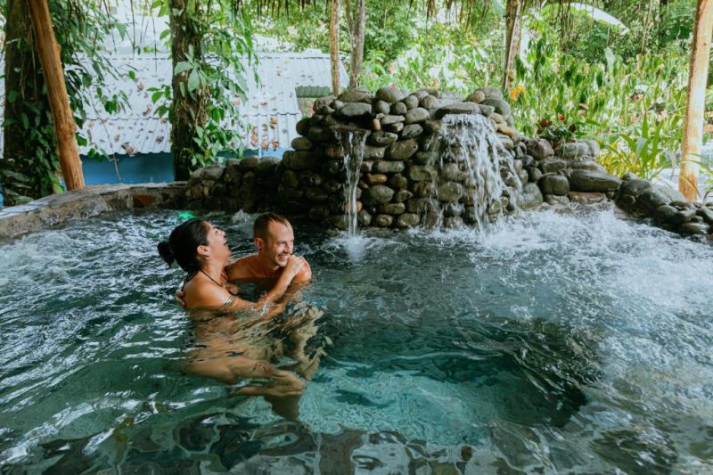 Couple enjoying a natural stone hot tub at a clothing-optional resort in Costa Rica, surrounded by lush greenery and tropical plants, creating a serene and relaxing atmosphere for naturists and adults.
