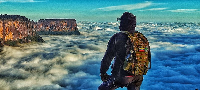 A person with a backpack stands on the edge of a cliff on Mount Roraima, overlooking a sea of clouds at sunrise. The rugged plateau is illuminated by golden sunlight, showcasing the breathtaking natural beauty of the sandstone formations. Perfect for adventure and helic
