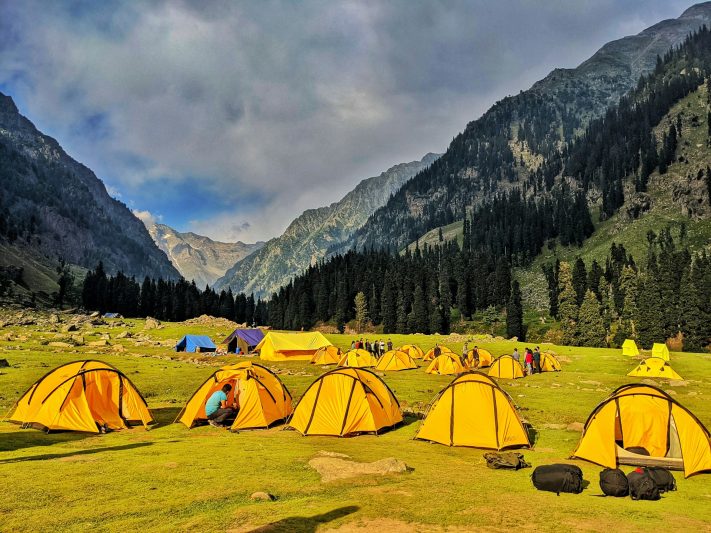 Bright yellow tents set up in a lush green valley surrounded by towering mountains, signifying a campsite during the Kashmir Great Lakes Trek.