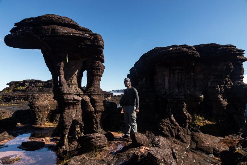 Tourist standing among unique rock formations on Mount Roraima during a helicopter tour, under a clear blue sky in 2025.
