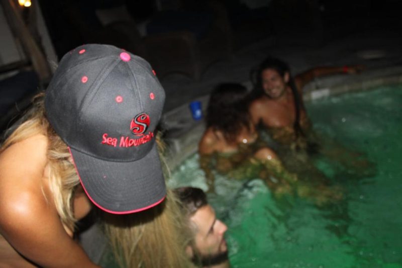 A group of people enjoying a night swim at a resort's pool, with one person wearing a cap featuring the logo 'Sea Mountain', capturing the relaxed and carefree atmosphere of a clothing optional vacation spot.
