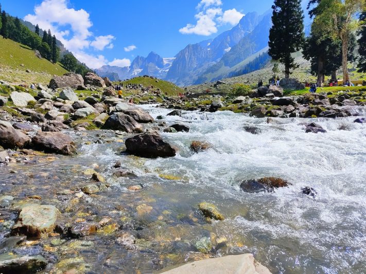 Scenic view of a flowing mountain stream surrounded by rocks and lush green landscape in Kashmir Great Lakes, under a bright blue sky with distant mountains in the background.