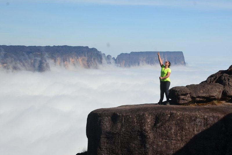 Person in bright clothing standing on a rocky cliff with arms raised, surrounded by a sea of clouds, with the majestic Mount Roraima in the background under a clear blue sky, illustrating the thrill of a helicopter tour