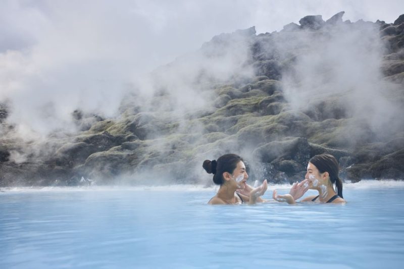 Two women enjoying a geothermal spa with steam rising, surrounded by rocky landscape in Iceland, highlighting relaxation and natural beauty.